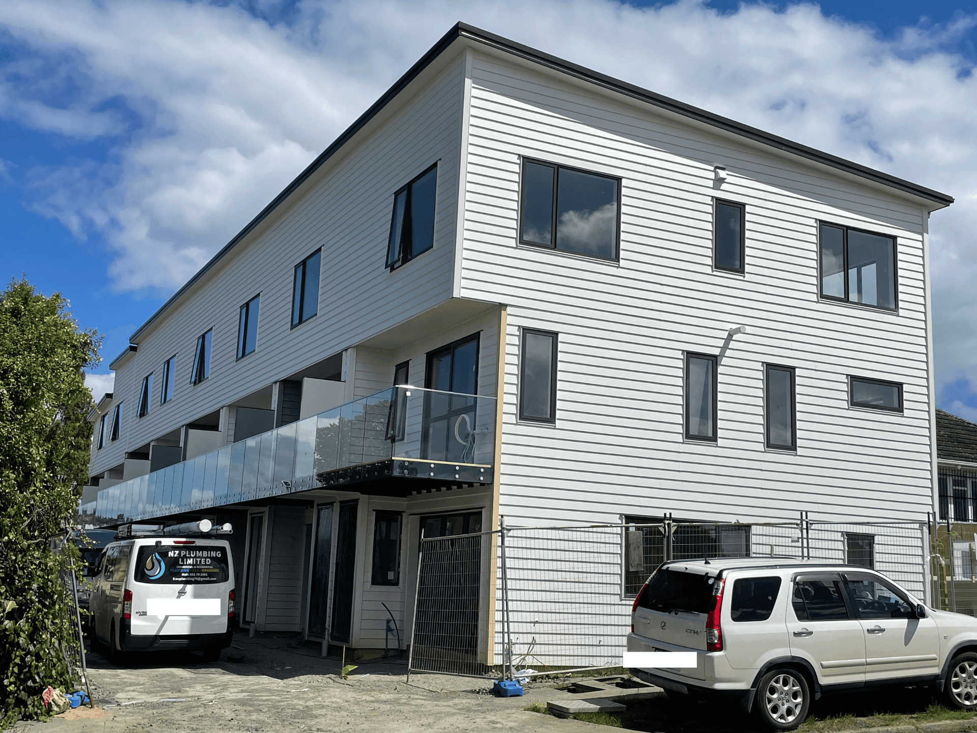 Modern three-story white building with multiple windows, glass balconies, and two parked vehicles in front.